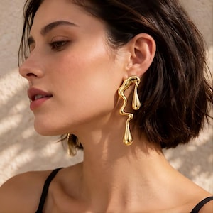 Puede incluir: Un par de pendientes dorados con un diseño abstracto y fluido de metal fundido. Los pendientes tienen una forma alargada e irregular con dos elementos en forma de gota que cuelgan de una estructura orgánica curva que cae hacia abajo.
