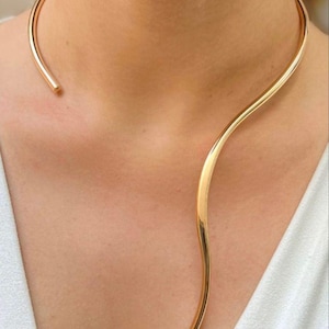 May include: A gold-toned metal choker necklace featuring a smooth, open-ended design. The piece consists of a single, continuous wire that curves around the neck, with one end resting near the collarbone and the other extending downward in a wave.