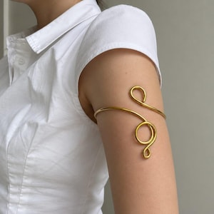 May include: A gold-coloured metal arm cuff featuring a continuous wire design that wraps around the upper arm. The piece has a smooth, polished finish and consists of two circular loops connected by a curved band, creating an open, adjustable shape.