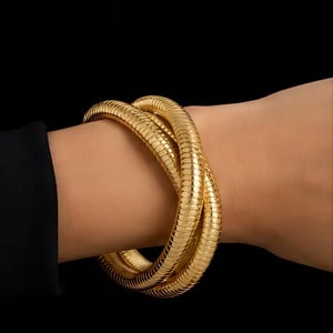 May include: A gold-toned bracelet featuring a flexible, segmented snake chain design that wraps around the wrist in a continuous, overlapping loop. The metal surface has a polished, reflective finish with a repeating, textured pattern throughout.