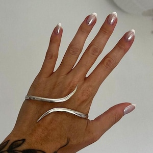 Op de afbeelding: Een zilverkleurige metalen manchetarmband met een bypass-ontwerp met twee taps toelopende, puntige uiteinden die om de pols buigen. De armband heeft een gladde, gepolijste afwerking en een open structuur die gedeeltelijk om de arm wikkelt.