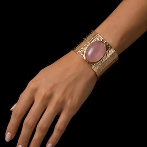 Puede incluir: Un brazalete ancho de tono dorado con una superficie de metal martillado y texturizado, que presenta una piedra grande, lisa y de forma ovalada en color rosa claro engastada en el centro. El brazalete tiene un acabado pulido y gran anchura.