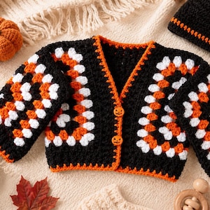 May include: A crochet cardigan featuring a black, orange, and white granny square pattern with long sleeves and two orange jack-o'-lantern buttons. The set includes a matching black crochet beanie with an orange pumpkin appliqué on the front.