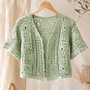 May include: A light green, open-front crochet cardigan with short sleeves. The cardigan features a floral pattern and a scalloped edge. It is displayed on a wooden hanger against a neutral background, showcasing the handmade details and open weave.