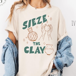 May include: An ivory t-shirt featuring the dark green text "SIEZE THE CLAY" alongside brown line art illustrations of two pottery vessels and a pair of hands holding a sphere, accented by small star shapes, worn with a blue denim jacket.