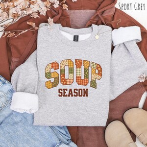 May include: A sport grey crewneck sweatshirt featuring the text SOUP SEASON in a block font. The letters are filled with a patchwork pattern of orange, green, and yellow floral and plaid designs. The word SOUP is arched, while SEASON is below.