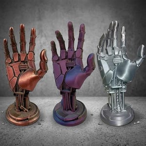 Könnte beinhalten: Drei metallische Roboterhand-Skulpturen in Kupfer, Lila und Silber. Jede Hand ist mit mechanischen Komponenten detailliert und auf einem kreisförmigen Sockel montiert. Die Hände sind in einer Geste der Begrüßung oder des Angebots dargestellt.