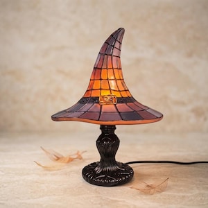 Peut inclure: Une lampe de table en vitrail en forme de chapeau de sorcière pointu. La lampe présente des panneaux de verre orange et violet disposés en grille, avec une base en métal sombre et un détail de boucle décorative sur la bande entourant la calotte.