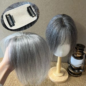 May include: A hair topper featuring straight, salt-and-pepper gray hair with a center part. The underside shows a beige base with two black metal pressure-sensitive clips for attachment. The hair is styled with soft bangs and a natural layered look.