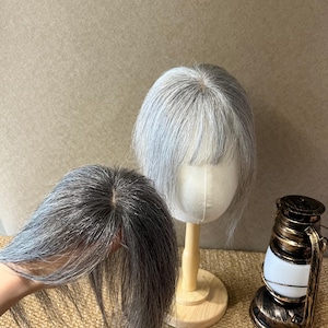 May include: A hair topper with straight, salt-and-pepper gray hair strands. One piece is displayed on a white mannequin head stand, while another identical hair topper is held in the foreground, showing the scalp-like base and hair texture.