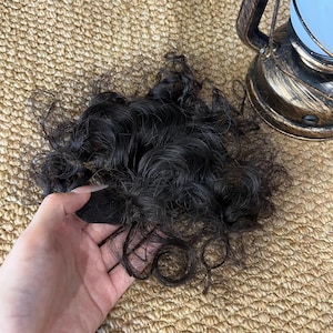 May include: A dark brown hair extension piece featuring tight, voluminous curls and a natural texture. The hair is gathered at a base, showcasing a dense arrangement of wavy strands that create a full, rounded shape with a deep, dark color profile.