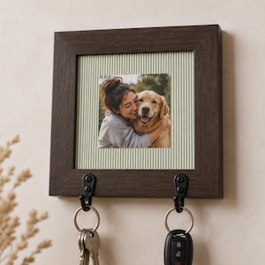 May include: A square dark brown wooden wall-mounted key holder featuring a central photo frame with a light green pinstriped mat. The frame displays a photograph of a person hugging a golden retriever. Two black metal hooks hold two key rings.