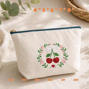 May include: A cream-colored canvas zipper pouch featuring an embroidered design of two red cherries hanging from a white bow, surrounded by a floral wreath. The text 4" 5" 6" 7" 8" and file formats HUS, VP3, EXP, PES, JEF, DST are visible.