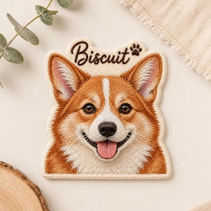 May include: An embroidered patch featuring the face of a brown and white Corgi dog with a pink tongue and the word Biscuit written in dark brown script above the head, accompanied by a small brown paw print icon.