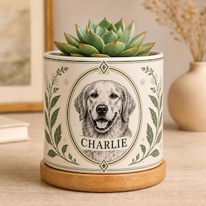 May include: A cylindrical ceramic planter with a cream-colored base and a wooden saucer. The planter features a green botanical border and a central oval frame containing a black and white illustration of a dog with the name CHARLIE below.