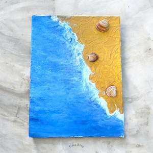 May include: A rectangular canvas art piece depicting a beach scene. The artwork features a blue ocean, golden sand, and three seashells. The text "Casa Riva" is visible at the bottom. The painting evokes a coastal theme.
