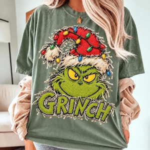 May include: A sage green t-shirt featuring a graphic of the Grinch wearing a red Santa hat adorned with colorful Christmas lights. The character has yellow eyes and a green face. The word GRINCH is printed in large green letters below.