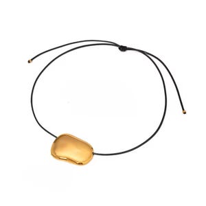 May include: A gold-colored pendant necklace with a black cord. The pendant has an irregular, organic shape. The necklace is displayed against a white background. The cord is adjustable.