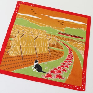 May include: A red fabric square with a colorful illustration of a rural landscape. The scene includes a field of hay bales, a winding path lined with red flowers, a train, and a black and white cat. The background features a sunset over a green hillside.