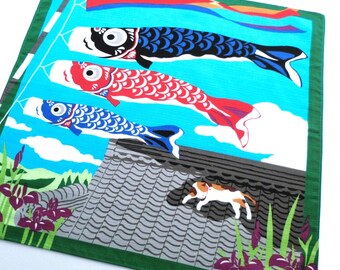 May include: A square fabric textile featuring a vibrant illustration of three colorful carp streamers flying against a bright blue sky. Below the streamers, a calico cat walks across a gray tiled roof, surrounded by green grass and purple iris flowers.
