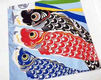 Può includere: Tessuto quadrato con tre colorati streamer a forma di carpa, un simbolo tradizionale giapponese. La carpa superiore è nera, quella centrale rossa e quella inferiore blu, tutte con motivi dettagliati. Lo sfondo è azzurro con strisce verdi e blu.