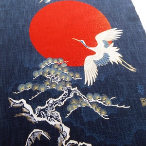 Puede incluir: Una tela azul con un diseño de inspiración japonesa que presenta dos grullas blancas volando sobre un sol rojo y un pino. La tela tiene una superficie texturizada y un diseño de color dorado.