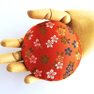 May include: A round, red fabric pincushion featuring a repeating pattern of small, multi-colored cherry blossom flowers in shades of white, pink, blue, and gold, with green leaves interspersed throughout the textile design.