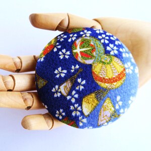 May include: A circular fabric-covered button featuring a deep blue background with a repeating pattern of white cherry blossoms, gold-accented folding fans, and spherical ornaments with red, green, and gold geometric details.