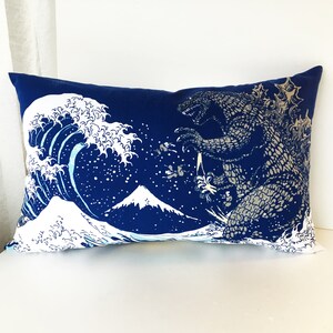 May include: A blue cushion featuring a white graphic of a wave, a mountain, and a Godzilla-like monster. The design is reminiscent of the famous Japanese woodblock print "The Great Wave off Kanagawa".