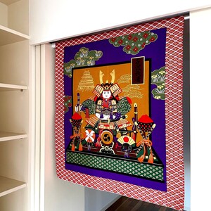 Könnte beinhalten: Eine violette und goldene Wandbehänge mit rotem Rand, die einen Samurai in traditioneller japanischer Kleidung zeigt. Die Wandbehänge hängt an einer weißen Wand.