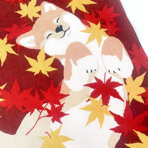 May include: A fabric textile featuring a tan and white Shiba Inu dog lying on its back with eyes closed, surrounded by a pattern of vibrant red and yellow maple leaves against a deep maroon background, creating a seasonal autumn-themed design.