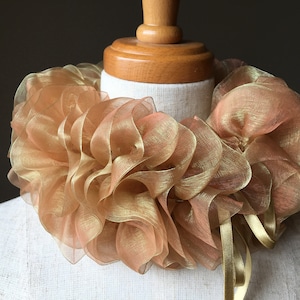 May include: A gold-colored, ruffled collar made of sheer fabric. The collar is displayed on a mannequin and features a layered design with ribbon ties. The fabric has a shimmering, metallic appearance.