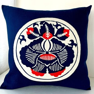 Op de afbeelding: Een donkerblauw kussen met een wit cirkelvormig ontwerp met een gestileerde rode en witte bloem. De bloem heeft een cirkelvormig midden en vijf bloemblaadjes.