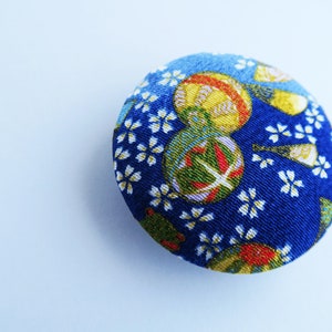 May include: A circular fabric-covered button featuring a vibrant blue background with a repeating pattern of small white cherry blossom flowers and larger, colorful spherical motifs in shades of yellow, green, and red with intricate gold detailing.