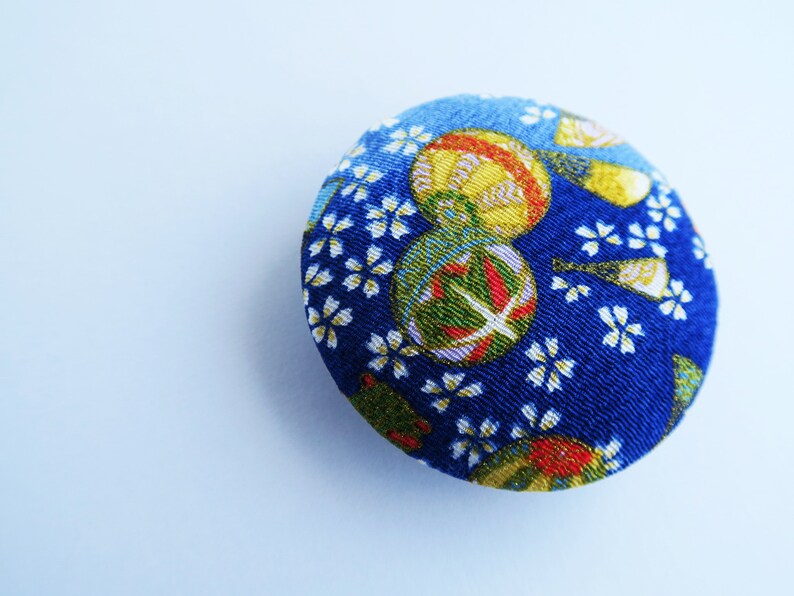 May include: A circular fabric-covered button featuring a vibrant blue background with a repeating pattern of small white cherry blossom flowers and larger, colorful spherical motifs in shades of yellow, green, and red with intricate gold detailing.