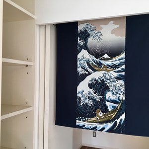 May include: A blue and white tapestry with a Japanese wave design. The wave is depicted in a traditional style with a mountain in the background. The tapestry is hanging on a white wall.