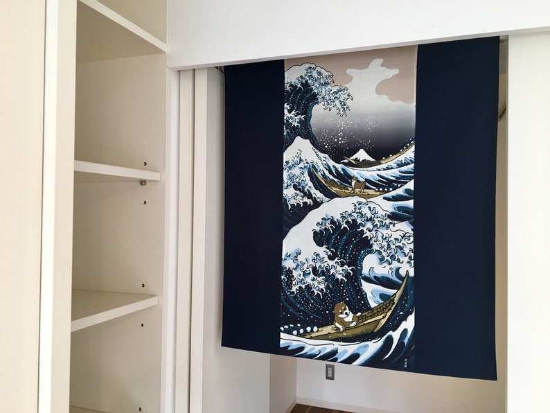 May include: A blue and white tapestry with a Japanese wave design. The wave is depicted in a traditional style with a mountain in the background. The tapestry is hanging on a white wall.