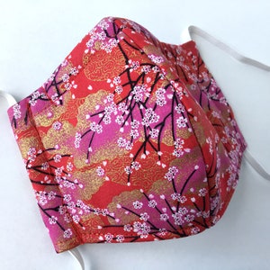 May include: A face mask featuring a vibrant red and pink fabric with a pattern of white cherry blossoms and gold cloud-like accents. The mask has a structured center seam and includes two white elastic ear loops attached to the sides for secure wear.
