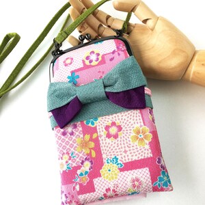 May include: A rectangular fabric pouch with a kiss-lock metal frame and a long green strap. The pouch features a pink background with a multi-colored floral pattern and a large, layered teal and purple fabric bow attached to the front.