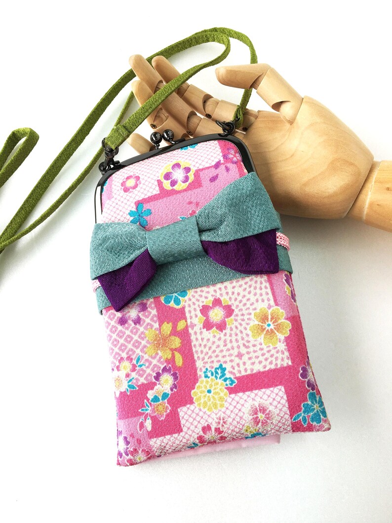 May include: A rectangular fabric pouch with a kiss-lock metal frame and a long green strap. The pouch features a pink background with a multi-colored floral pattern and a large, layered teal and purple fabric bow attached to the front.