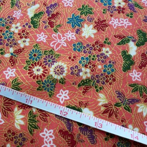 May include: A close-up of a coral pink fabric with a repeating pattern of gold, green, blue, and purple flowers and butterflies. A white measuring tape is laid diagonally across the fabric, showing measurements in inches.
