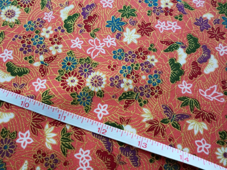 May include: A close-up of a coral pink fabric with a repeating pattern of gold, green, blue, and purple flowers and butterflies. A white measuring tape is laid diagonally across the fabric, showing measurements in inches.