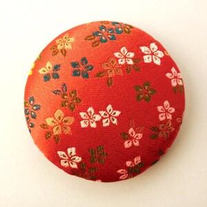 May include: A circular fabric-covered button featuring a vibrant red background with a repeating pattern of small, multi-colored floral blossoms in shades of white, light pink, gold, and teal, with green leaves interspersed throughout the design.