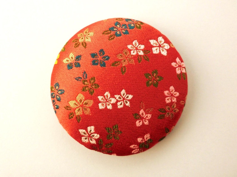 May include: A circular fabric-covered button featuring a vibrant red background with a repeating pattern of small, multi-colored floral blossoms in shades of white, light pink, gold, and teal, with green leaves interspersed throughout the design.