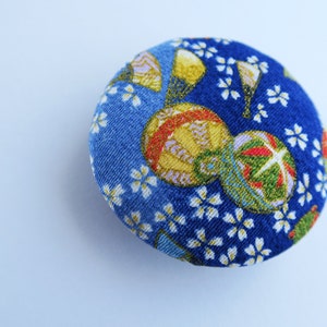 May include: A circular fabric-covered button featuring a vibrant blue background with a repeating pattern of small white cherry blossoms and larger, detailed gold and green decorative spheres with intricate line work and metallic accents.