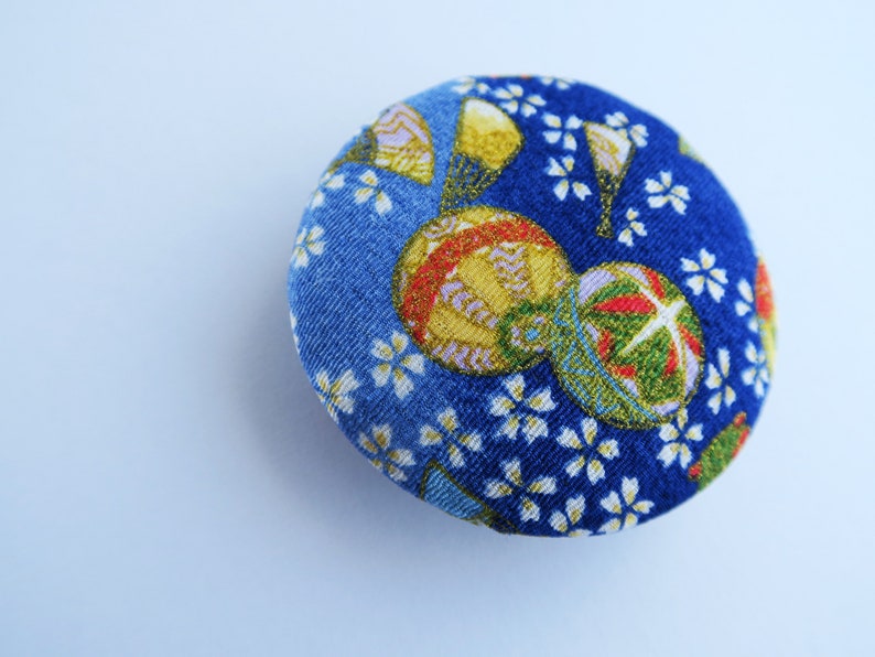 May include: A circular fabric-covered button featuring a vibrant blue background with a repeating pattern of small white cherry blossoms and larger, detailed gold and green decorative spheres with intricate line work and metallic accents.