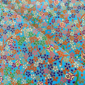 Puede incluir: Una tela de color azul brillante con un patr&oacute;n denso y repetitivo de peque&ntilde;as flores en tonos naranja, blanco, morado y verde, intercaladas con motivos circulares dorados y finas ramas marrones entrelazadas por toda la superficie.