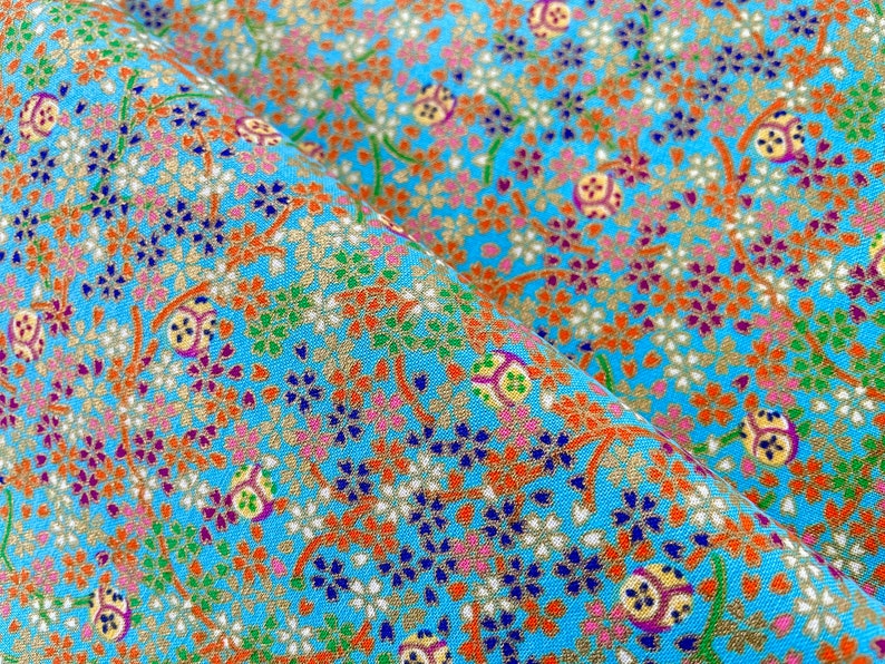 Puede incluir: Una tela de color azul brillante con un patr&oacute;n denso y repetitivo de peque&ntilde;as flores en tonos naranja, blanco, morado y verde, intercaladas con motivos circulares dorados y finas ramas marrones entrelazadas por toda la superficie.