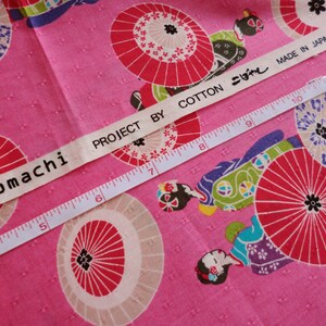 May include: Pink fabric with a white label that reads "komachi PROJECT BY COTTON こぼし MADE IN JAPAN". The fabric has a repeating pattern of red and white umbrellas and women in traditional Japanese clothing.