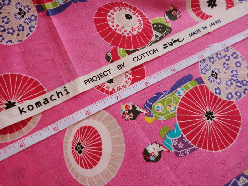 May include: Pink fabric with a white label that reads "komachi PROJECT BY COTTON こぼし MADE IN JAPAN". The fabric has a repeating pattern of red and white umbrellas and women in traditional Japanese clothing.